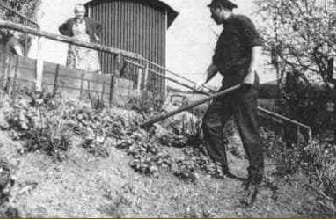 Ein Mann mit Hut bearbeitet mit einer Hacke einen abschüssigen Garten. Eine Frau steht hinter einem niedrigen Zaun und schaut zu. Im Hintergrund ist ein Holzschuppen zu sehen, und rechts ist ein Baum mit Blättern zu sehen. Die Szene ist in Schwarzweiß gehalten.