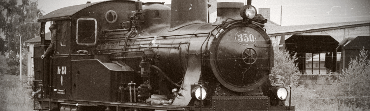 Ein altes Schwarzweißfoto einer Dampflokomotive mit der Nummer 350 auf der Vorderseite. Die Lokomotive steht im Freien, im Hintergrund sind Teile von Gebäuden zu sehen, umgeben von Vegetation.